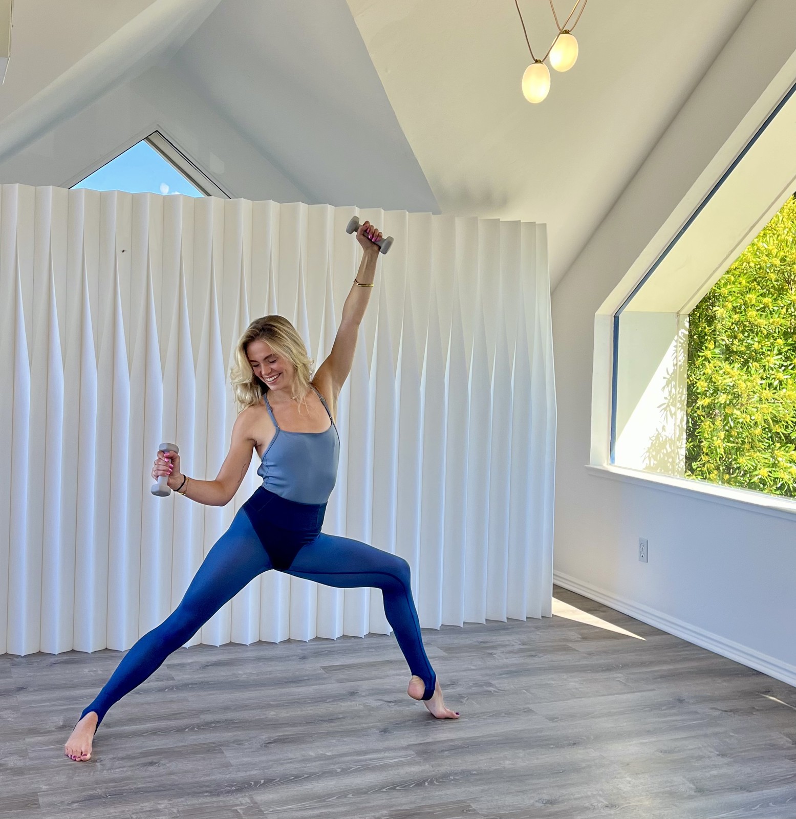 Lily in a wide standing stance holding two small hand weights in a sunlit studio