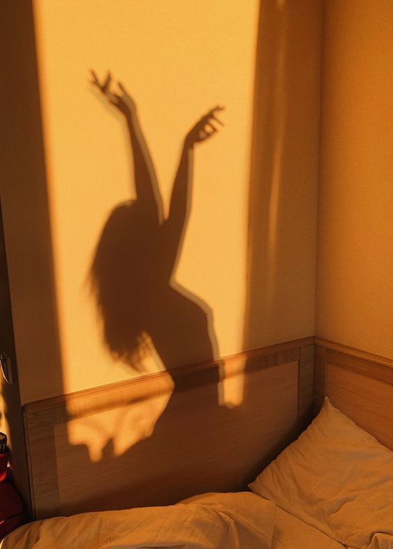 The shadow of a dancer with raised arms cast across a golden-lit bedroom wall